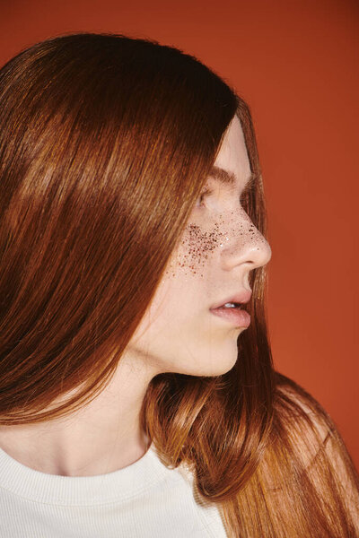 A young man with long red hair displays his fashionable style against a warm, vibrant backdrop.