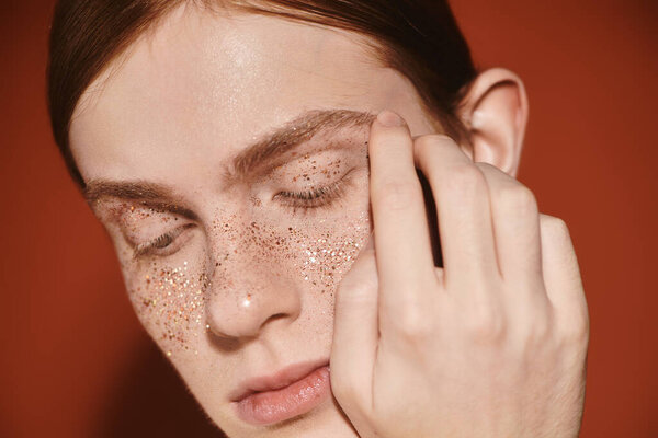 A young queer man showcases his elegant style with shimmering glitter on his face in a cozy setting.