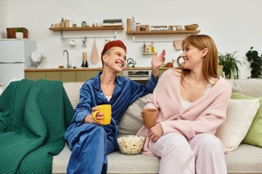 A lovely lesbian couple laughs and shares popcorn in their modern home. clipart