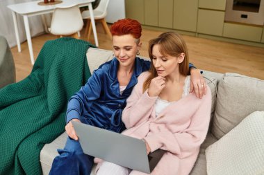 Two beautiful women relax at home, sharing laughter while using a laptop on the sofa. clipart