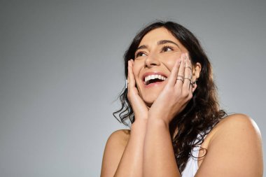 Gri arka planda beyaz atletli, iri yarı, gülümseyen genç bir kadın..