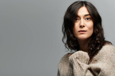 Esmer, yumuşak hatlı bir kadın gri bir arka planda günlük giysiler içinde poz veriyor..