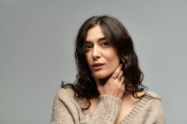 Esmer saçlı genç bir kadın gri bir arka planla stüdyoda zarifçe poz veriyor..