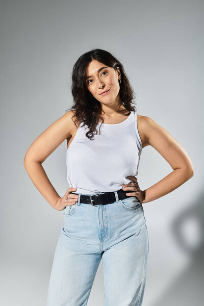 Young plus size woman confidently poses in white tank top against grey backdrop.