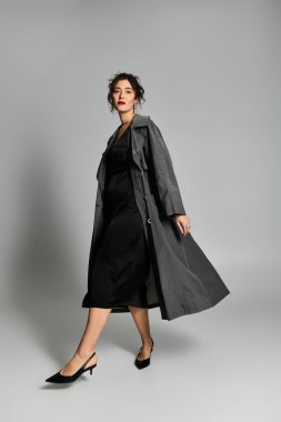 Siyah elbiseli ve gri ceketli genç bir kadın stüdyoda zarif pozlar veriyor..