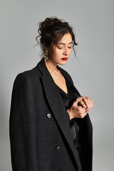 Брюнетка в элегантном черном наряде изящно позирует на сером фоне.