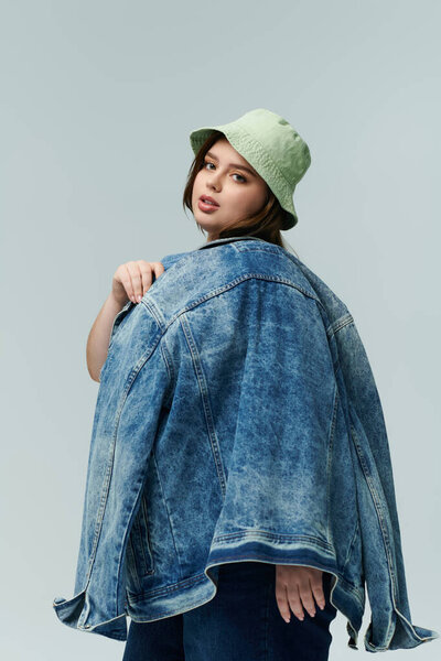 Against a simple backdrop, a beautiful young woman models a denim jacket with a trendy bucket hat.