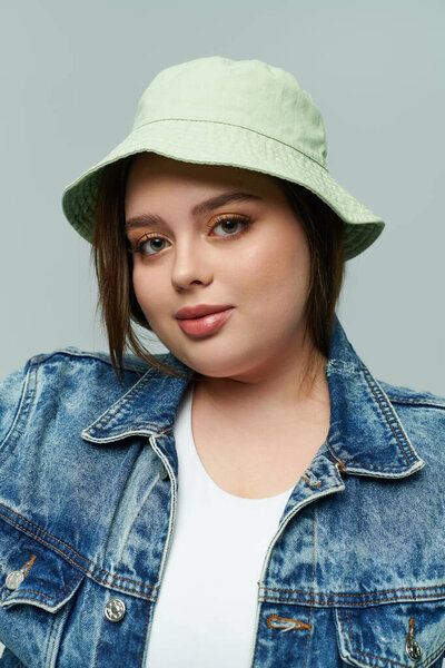 Confident plus size woman in a denim jacket and light bucket hat poses against a neutral backdrop.