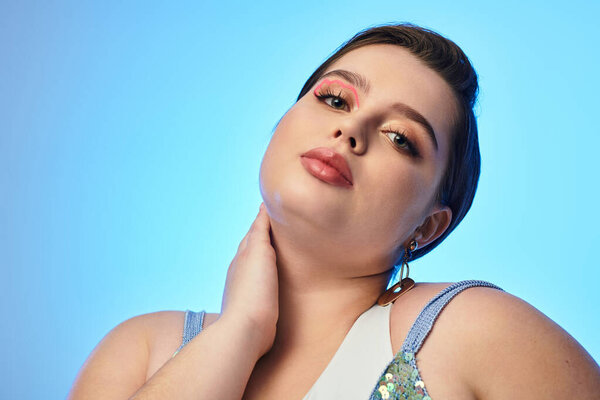 Plus size woman with striking makeup poses elegantly under soft blue lighting, embracing her beauty.