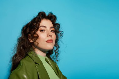 Kıvırcık saçlı, kendine güvenen genç bir kadın parlak mavi bir arka plana karşı duruyor..