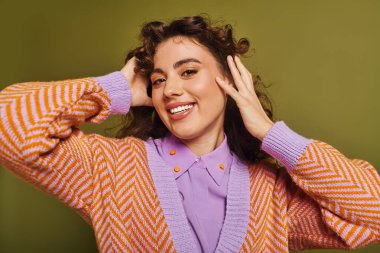 Kıvırcık saçlı, genç ve şık bir kadın, parlak yeşil bir duvara karşı neşe içinde poz verirken gülümser..