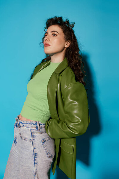 Молодая женщина в зеленом топе и куртке элегантно стоит, излучая игривую уверенность.