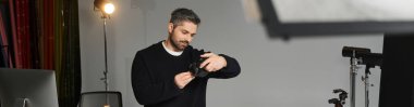 A skilled man adjusts his camera gear while preparing for a session in a well lit studio. clipart