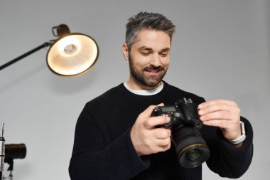 A man with a beard is focused on adjusting his camera while in a well lit studio. clipart