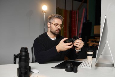 A man examines his camera settings while seated at a stylish desk in a contemporary studio. clipart