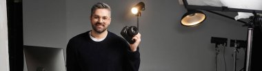 Adam, iyi donanımlı bir fotoğraf stüdyosunda elinde kamerayla gülümsüyor..