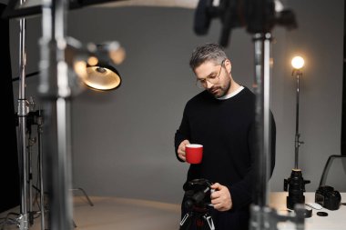 A man enjoying a warm beverage while setting up his camera in a well lit studio environment. clipart