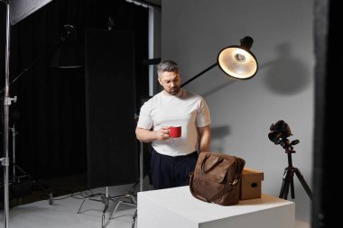 A man stands in a well lit studio holding a red cup of coffee, ready for his next project. clipart