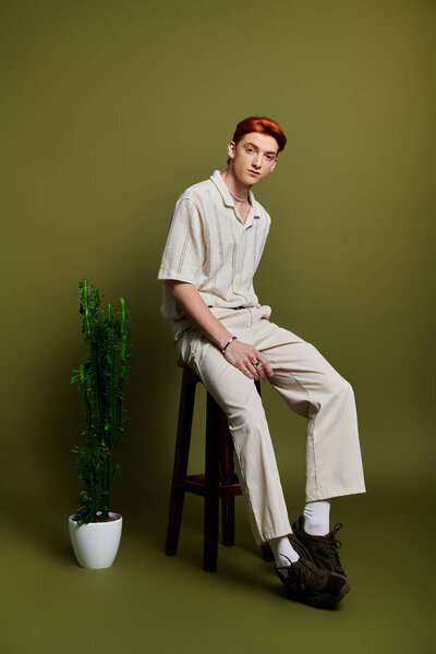 A thoughtful young man sits on a stool among calming greens and a potted plant.