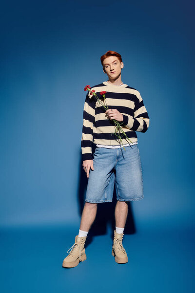A handsome young man stands confidently, holding roses, wearing a striped sweater and denim shorts.