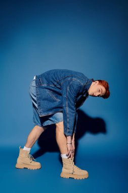 Genç bir adam çizmesini ayarlıyor, benzersiz modasını mavi bir arka plana vurguluyor..