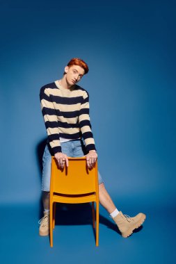 Çarpıcı özellikleri olan duygusal genç bir adam renkli bir sandalyenin üzerine eğilip tarzını sergiliyor..