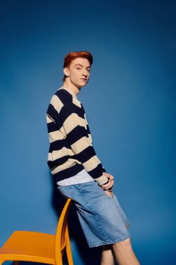 Genç, duygusal bir adam, mavi bir arka planla, düşüncelere dalmış, gündelik bir şekilde sandalyede oturuyor..