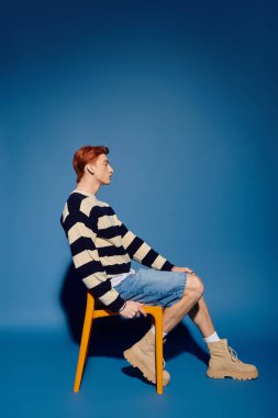 Genç bir adam düşünceli bir şekilde canlı bir sandalyede oturur, derin duygularını mavi bir ortamda ifade eder..