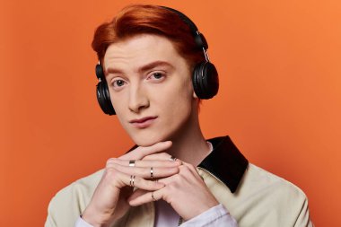 A young and handsome man contemplates while wearing headphones, framed by a bright orange wall. clipart