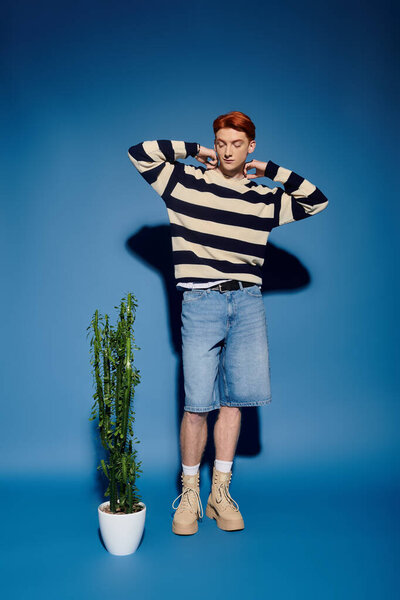 A young man with striking features shifts near a potted plant, lost in deep thought.