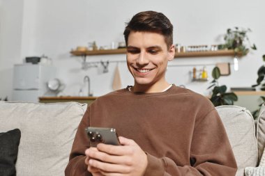 Kanepede akıllı telefonunu kontrol eden genç bir adam gülümserken evde rahat bir atmosfer var..
