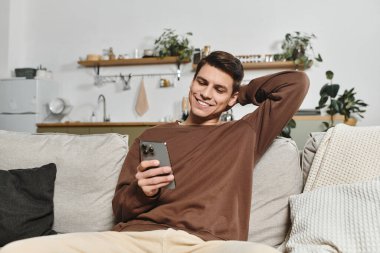 Genç bir adam rahat bir şekilde yumuşak bir koltukta oturuyor, akıllı telefonuyla meşgul, gülümsüyor.
