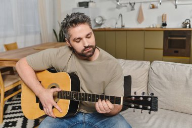 Bir adam yumuşak bir koltukta otururken gitar çalar, sade ve rahatlatıcı bir atmosfer yaratır..