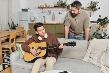 Genç bir adam rahat bir kanepede gitar çalarken bir aile üyesi neşeyle izliyor..