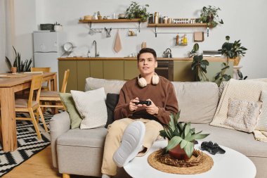 A young man sits on a soft sofa, fully engaged in playing video games in a warm, inviting home. clipart