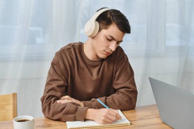 Genç adam sıcak, davetkar bir iç mekanda ahşap bir masada notlar alırken müzik dinliyor..