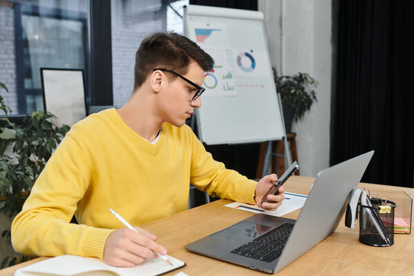 Новый сотрудник сидит за столом, сосредоточенный на смартфоне, работая над ноутбуком в офисе.