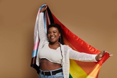 A joyful woman in trendy outfit holding pride flag and smiling cheerfully. clipart