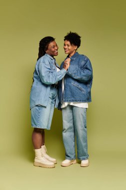 İki modaya uygun kadın, canlı yeşil bir arka planda şık kot elbiselerle aşkı kutluyor..