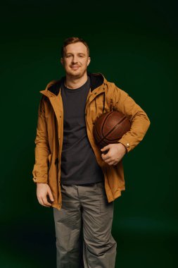 Sıradan kıyafetler giyen yakışıklı bir adam, iyi aydınlatılmış bir stüdyoda basketbol topu tutuyor..