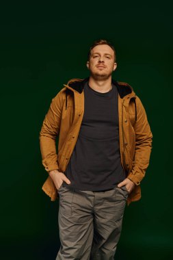 Kendine güvenen, sıradan giyinen yakışıklı bir adam elleri ceplerinde yeşil bir arka planda duruyor..