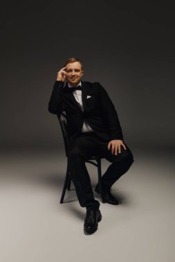 Yakışıklı bir adam, smokin ve papyon giymiş, kendine güvenen bir şekilde stüdyoda oturuyor..