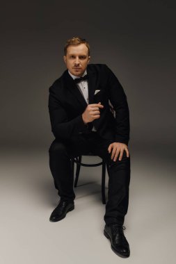 Smokinli şık bir adam, kendine güvenen bir şekilde sandalyeye oturur ve duruşunu belli eder..