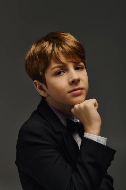 Smokin ve papyon giymiş genç bir çocuk stüdyoda şık bir poz sergiliyor..