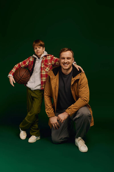 Счастливый отец преклоняет колени рядом со своим сыном с баскетбольным мячом, оба в стильной повседневной одежде.