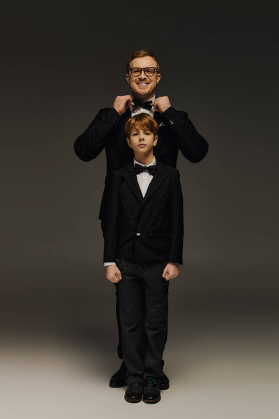 Красивый отец настраивает бабочку своего сына, демонстрируя их одинаковые смокинги в студии.