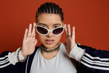 Kendine güvenen genç bir kadın şık güneş gözlükleri ve karmaşık örgüleriyle poz veriyor..