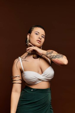 Kendine güvenen, modaya uygun giyinmiş genç bir kadın kahverengi bir arkaplanda zarif bir şekilde poz veriyor..