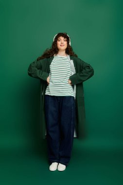 Kendine güvenen genç bir kadın, elleri kalçasında poz verir, yeşil bir ortamda şık elbisesiyle hava atar..