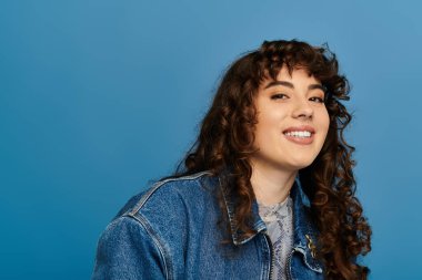 Young woman with curly hair wears a denim jacket and radiates happiness against a blue backdrop. clipart